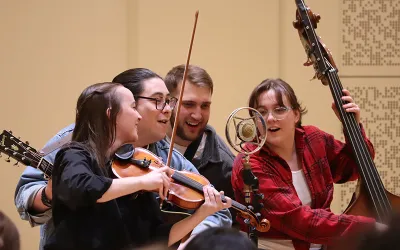 Students with a fiddle, string bass and guitar singing into a microphone.