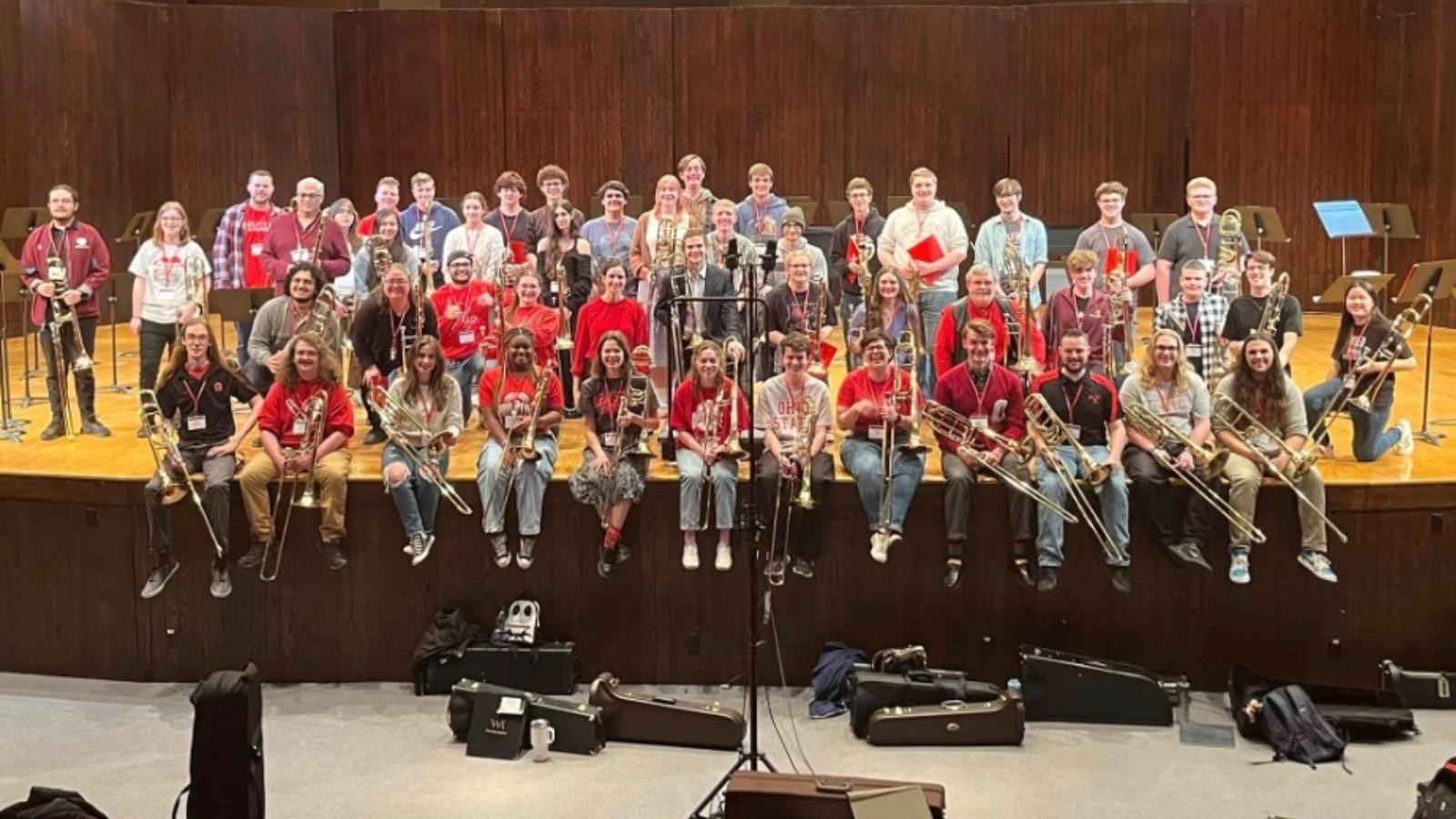 50 students holding trombones sitting on front edge of a stage