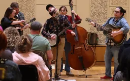 Students with folk-style string instruments playing for a seated audience.
